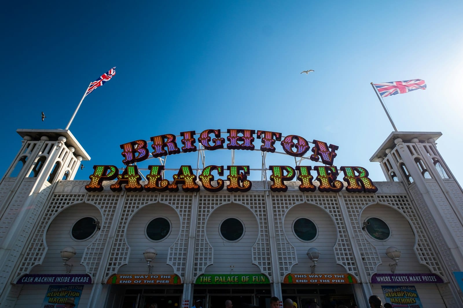 Ingresso del Brighton Palace Pier con bandiere britanniche, cielo azzurro limpido e persone che passano sotto un cartello colorato in una giornata di sole