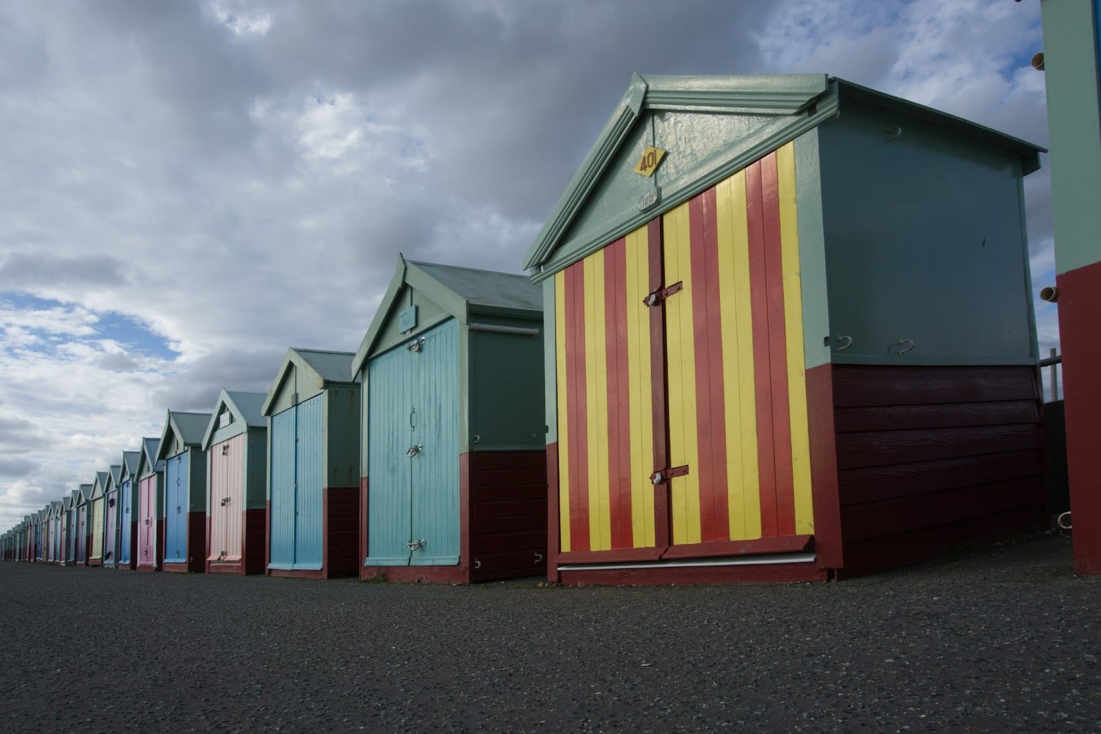 Reihe ikonischer pastellfarbener Strandhütten an der Strandpromenade von Brighton unter einem dramatisch bewölkten Himmel, ohne weitere nennenswerte Merkmale.