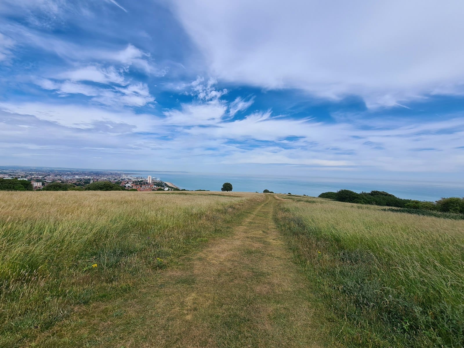 Breiter Grasweg auf einem Hügel mit Blick auf das Meer und die Stadt Brighton unter einem blauen Himmel mit vereinzelten Wolken.