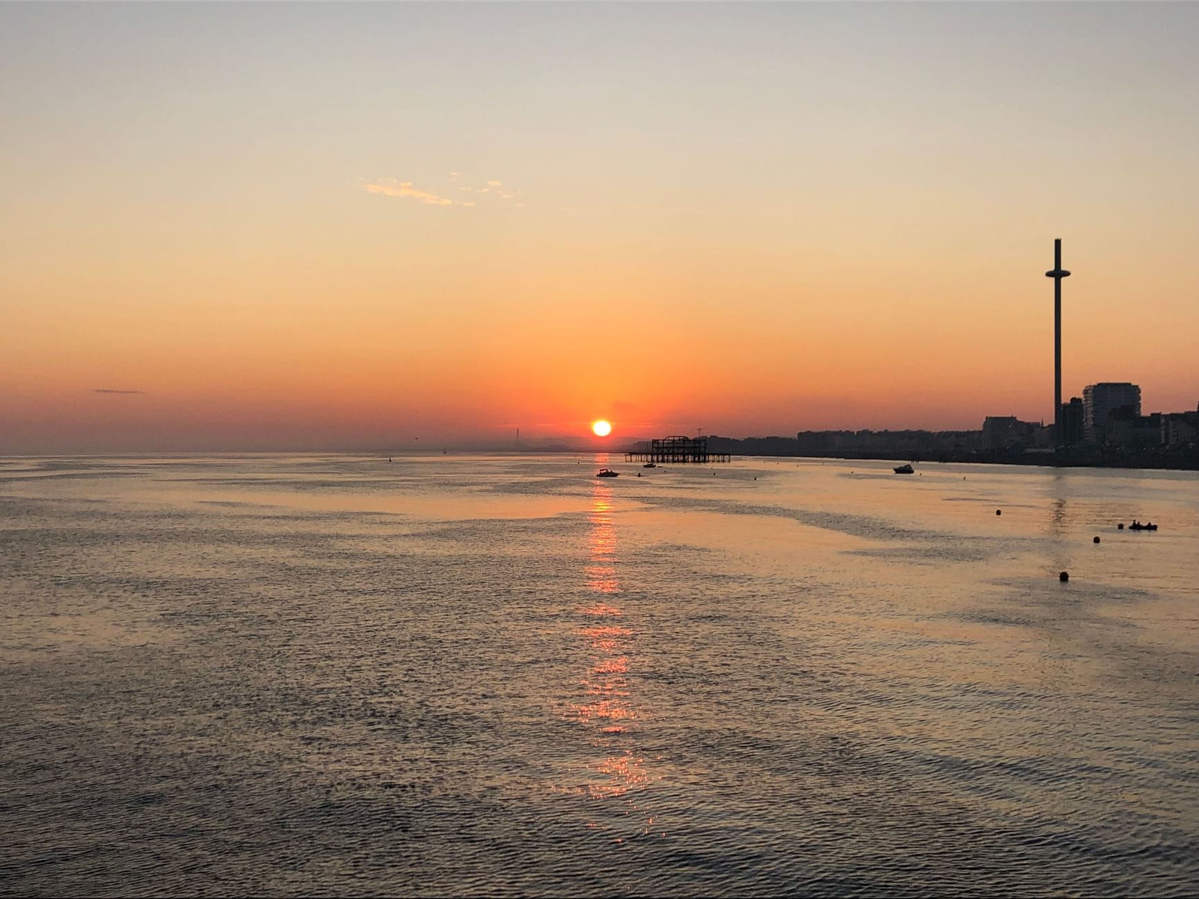 Brighton i360 observation tower silhouetted against an orange sunset sky, with calm sea and distant pier on Brighton seafront.