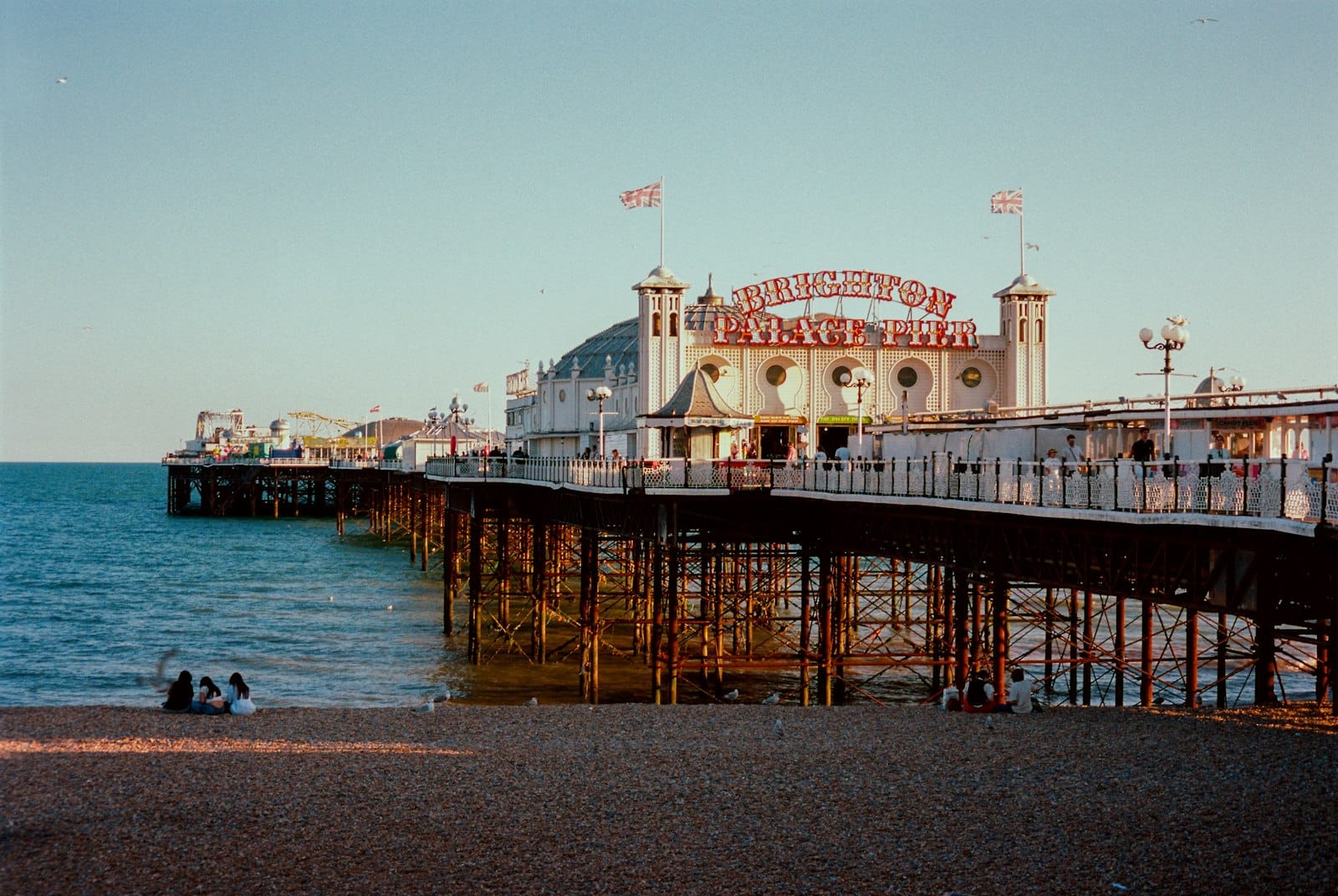 Der Brighton Palace Pier erstreckt sich über das Meer, im Vordergrund entspannen Menschen am Kieselstrand, unter einem blauen Sommerhimmel.