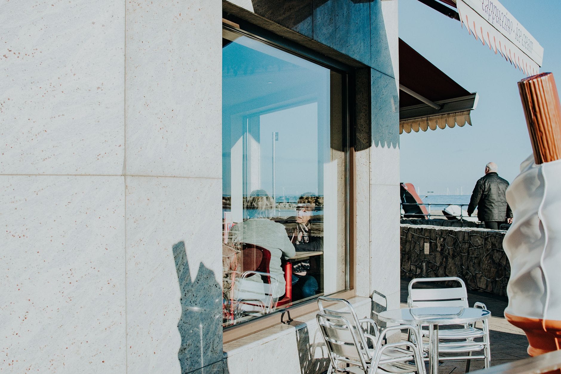 Außenbereich eines Strandcafés in Brighton mit Metallstühlen und Spiegelungen von Menschen, die in der Sommersonne Getränke genießen.