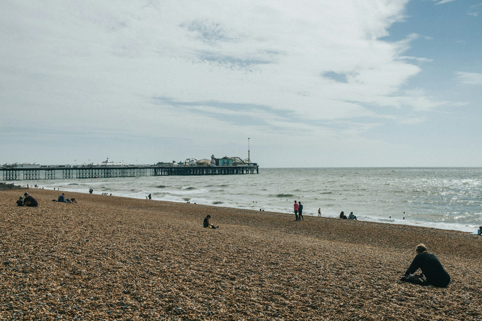 Kieselstrand in Brighton mit einigen sitzenden Menschen, Brighton Pier im Hintergrund und einem windigen Himmel.