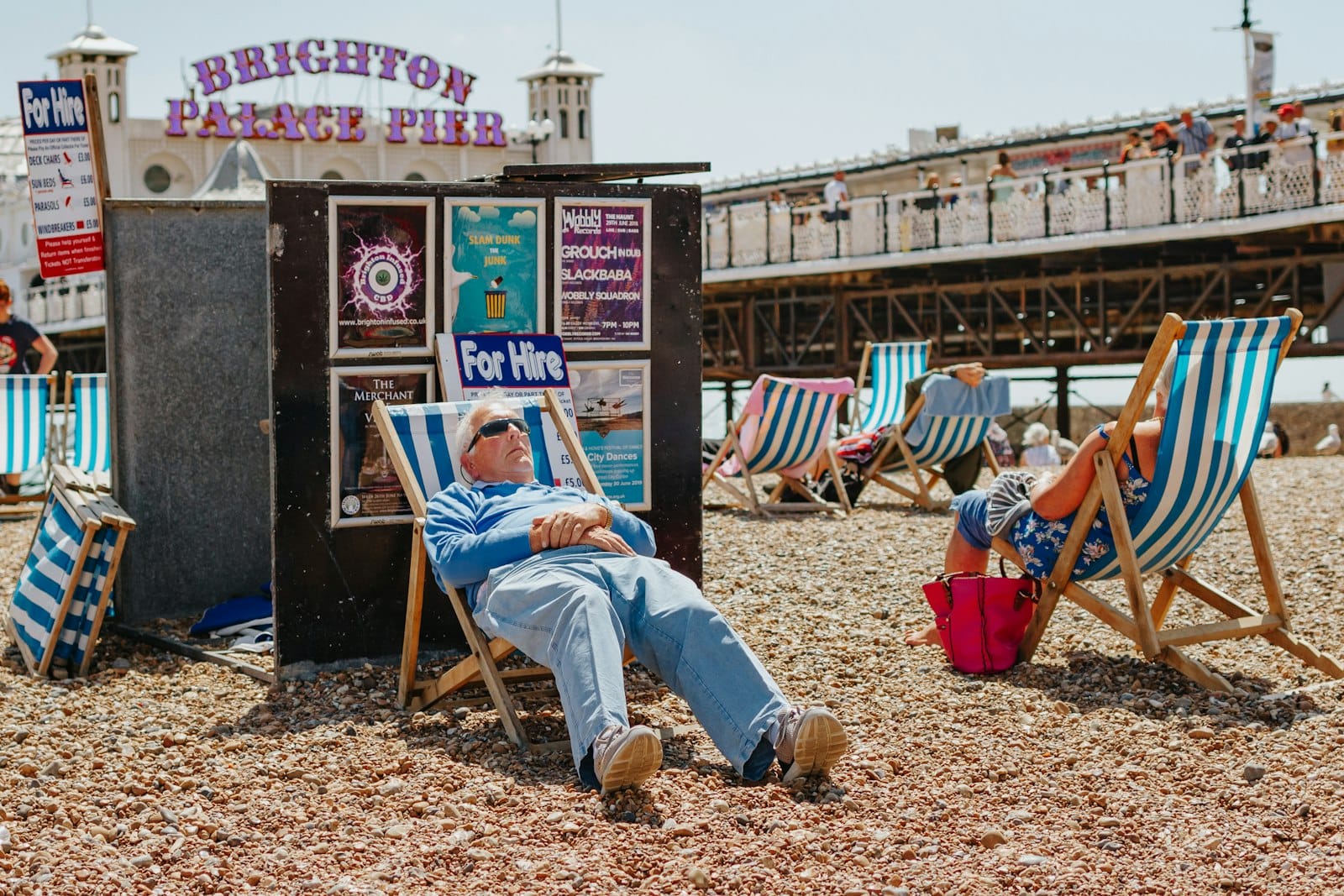 People relaxing on Brighton beach in deckchairs with Brighton Palace Pier in the background under sunny summer skies.