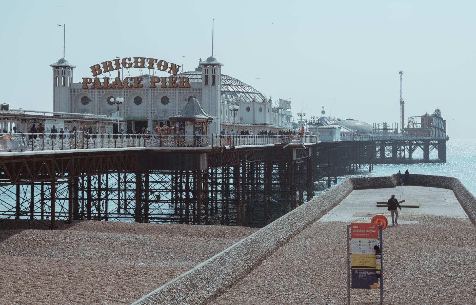 Der Brighton Palace Pier erstreckt sich über einen Kieselstrand mit einer breiten Strandpromenade, ein paar Menschen spazieren an einem kühlen, dunstigen Tag im Freien.
