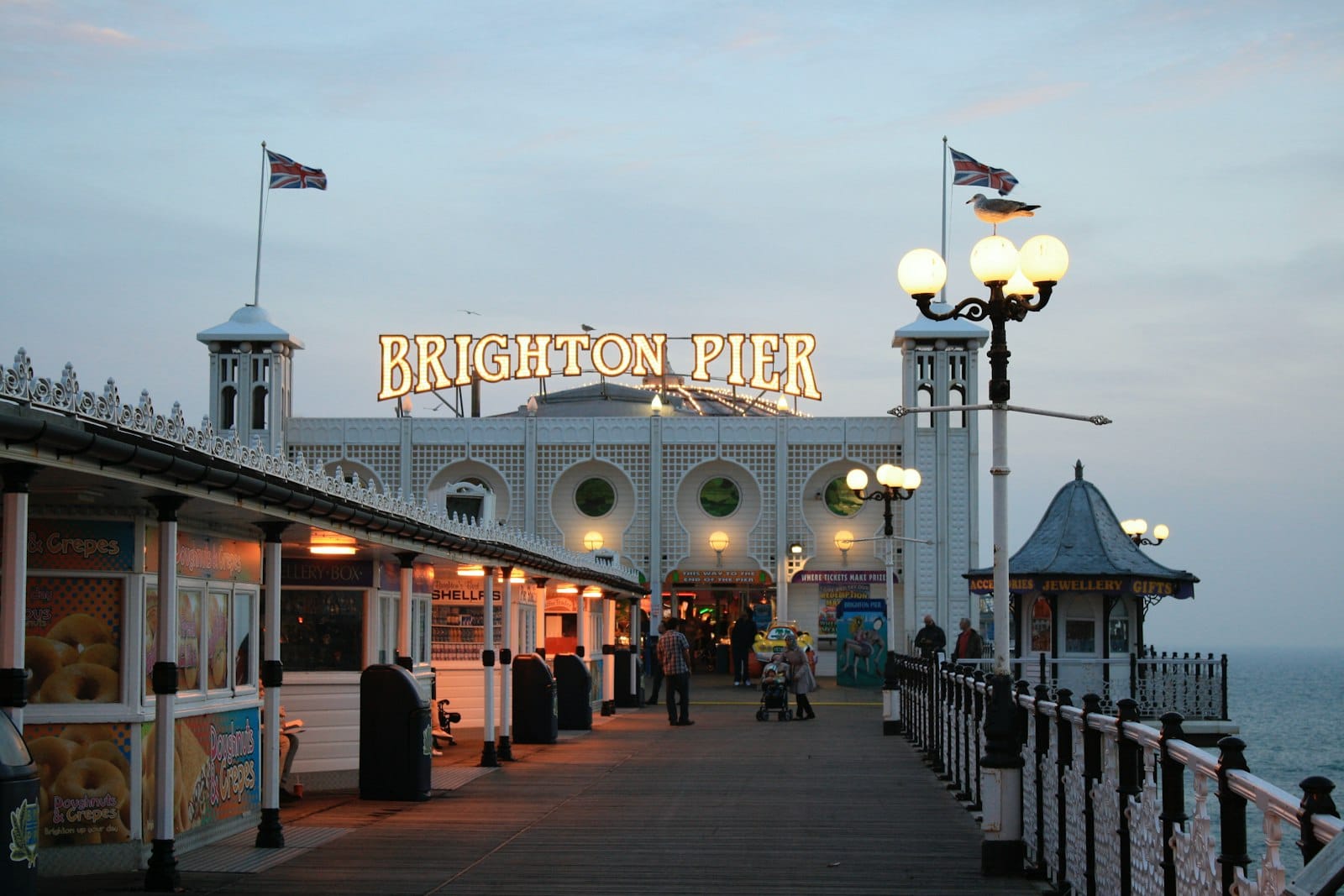View along Brighton Pier at dusk, people strolling, illuminated lights and iconic sign overhead, with seaside atmosphere and overcast sky.