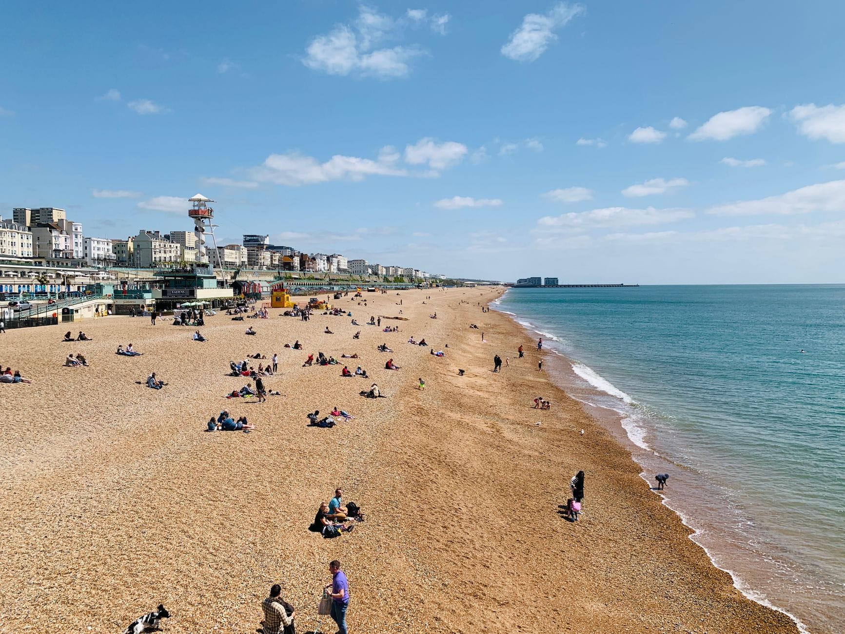 Szeroki widok na plażę Brighton z rozproszonymi odwiedzającymi pod błękitnym niebem, ukazujący linię brzegową i miasto w tle.