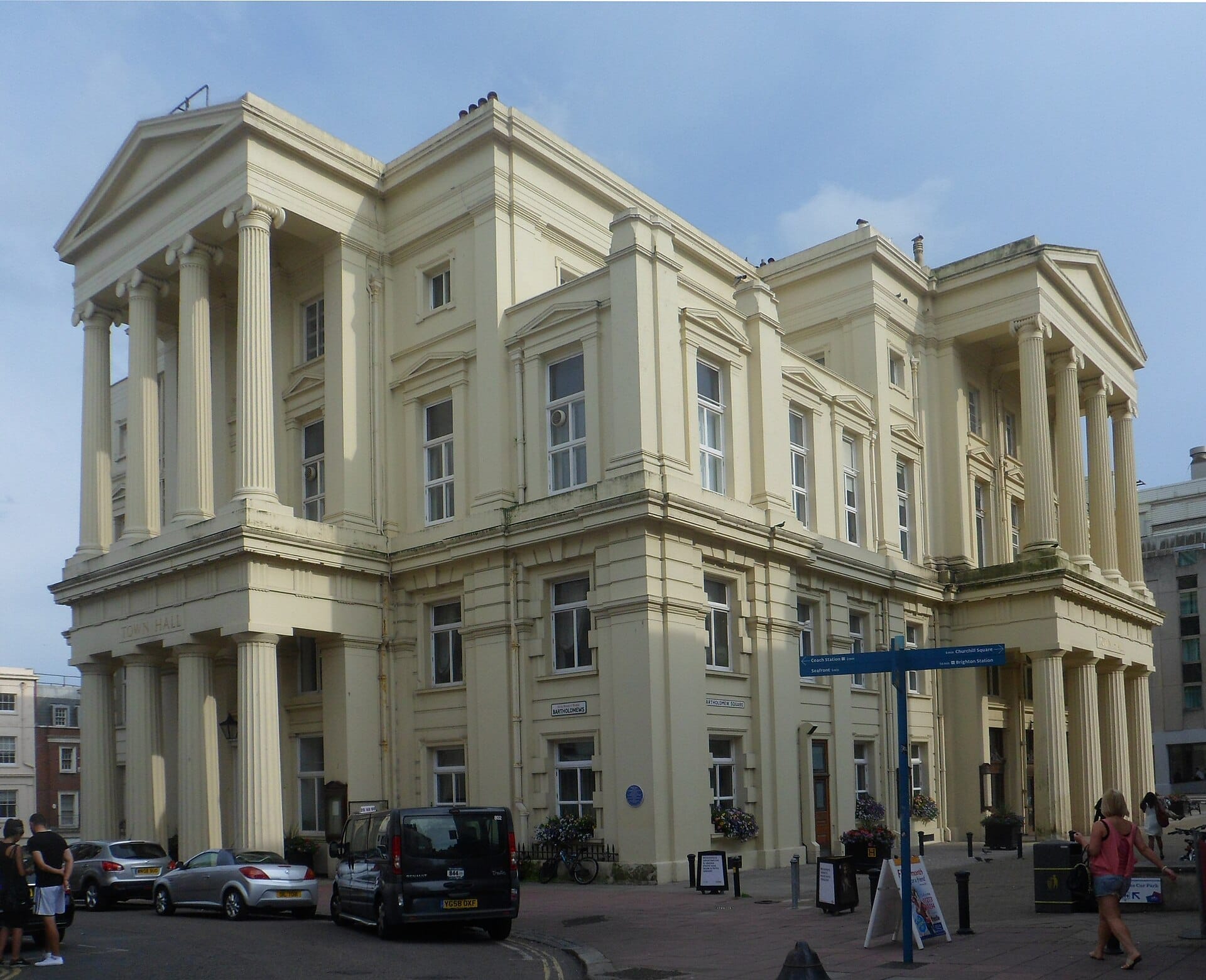 Brighton Town Hall exterior with grand neoclassical columns, pale yellow facade, parked cars, and people on the street under a clear sky.