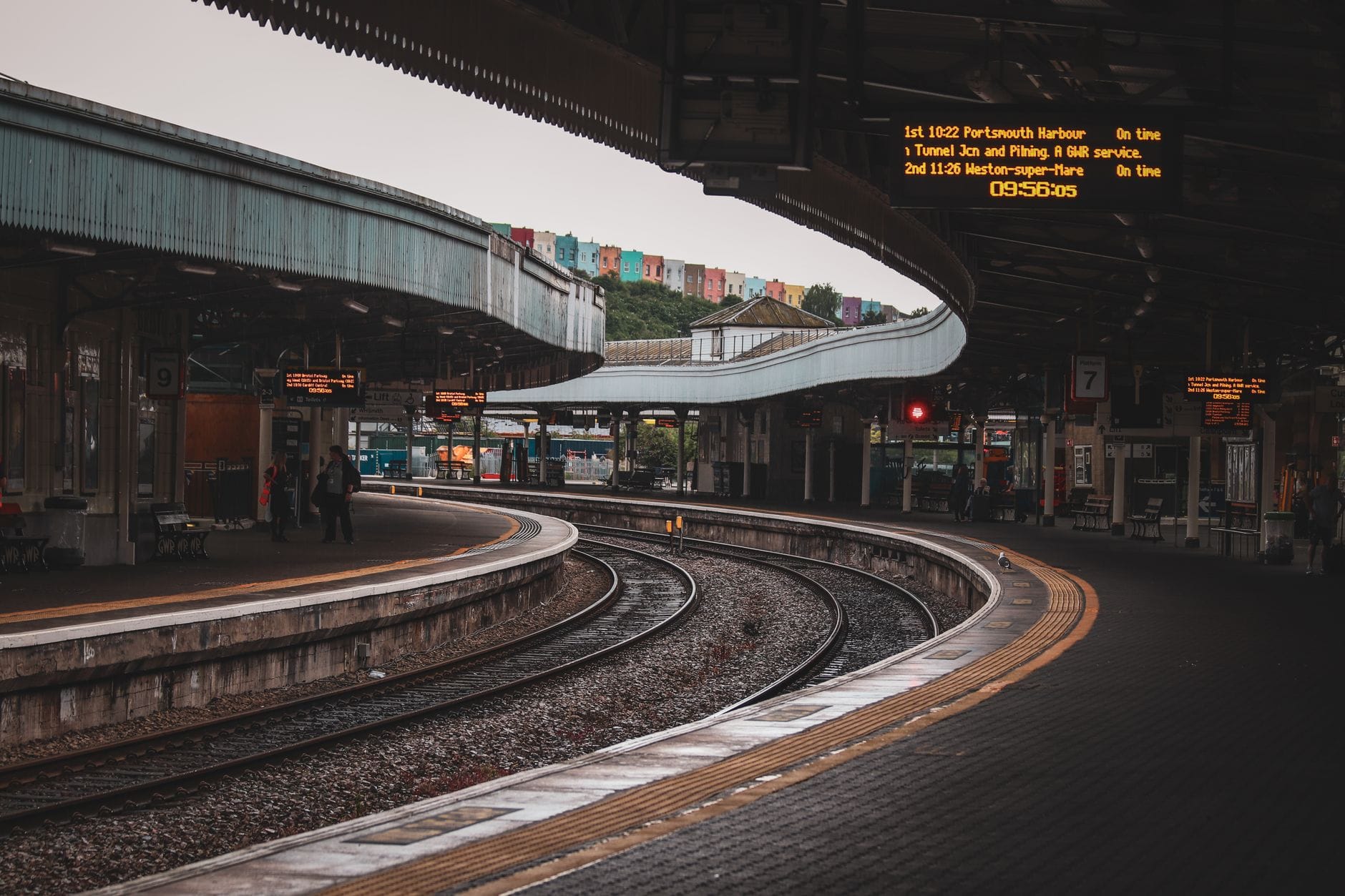 Andenes curvos y vías en la estación de tren de Brighton con paneles digitales de salidas y casas coloridas al fondo.