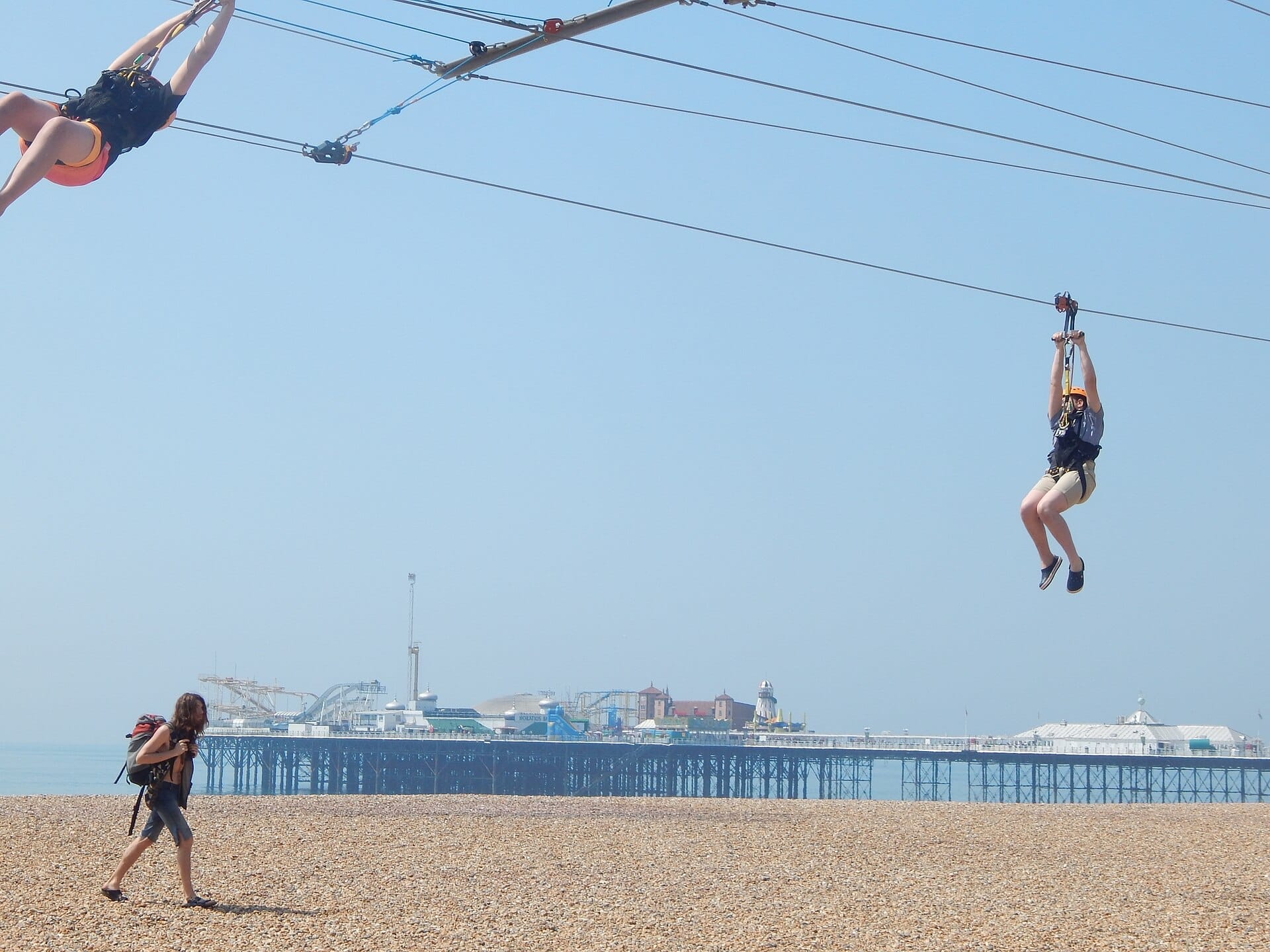 Two people ride the Brighton Zip wire above the pebbly beach, with Brighton Palace Pier visible in the background on a clear day.