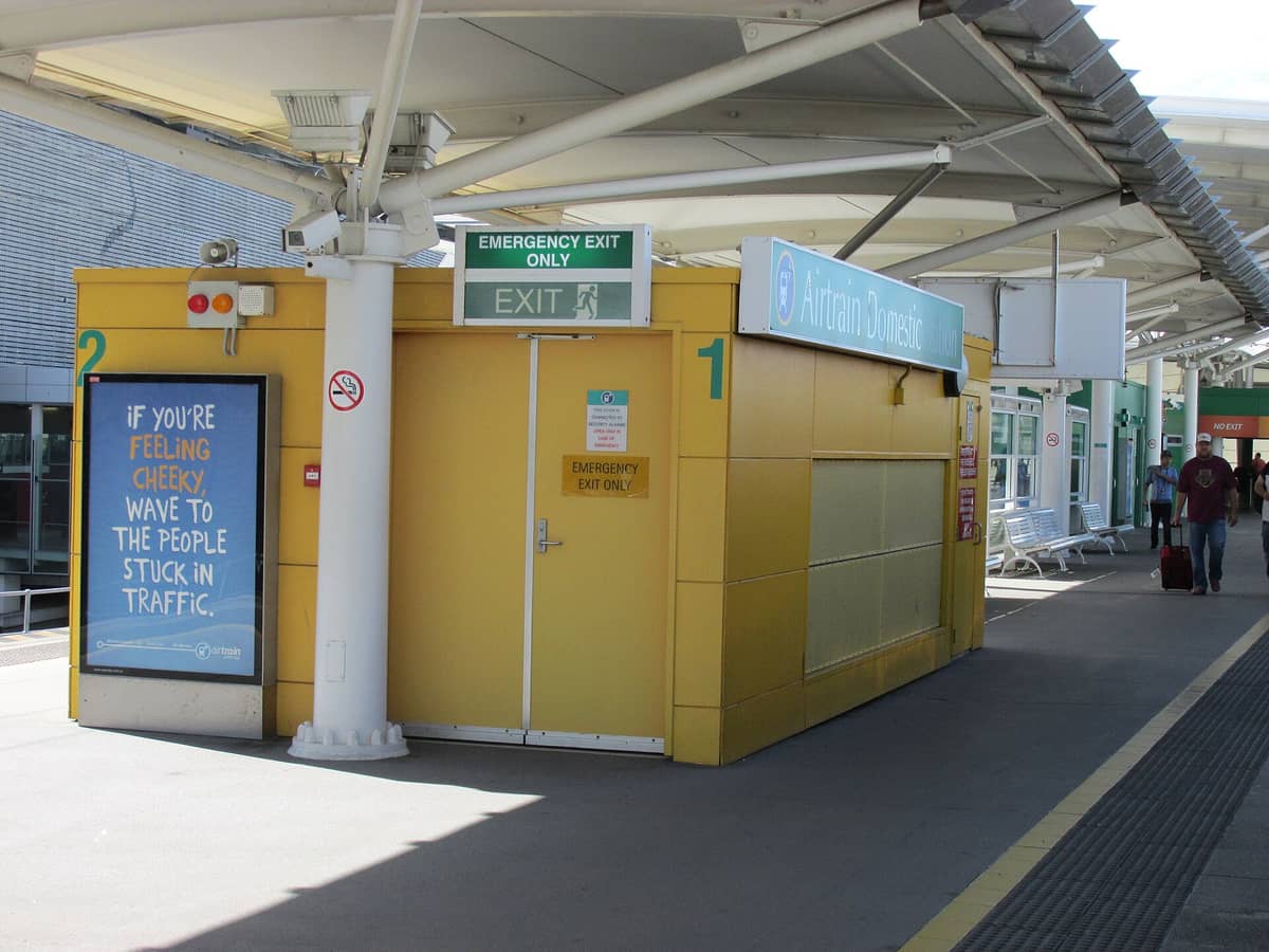 Airtrain Domestic Station at Brisbane Airport with yellow walls, signage, covered walkway, and people walking with luggage.