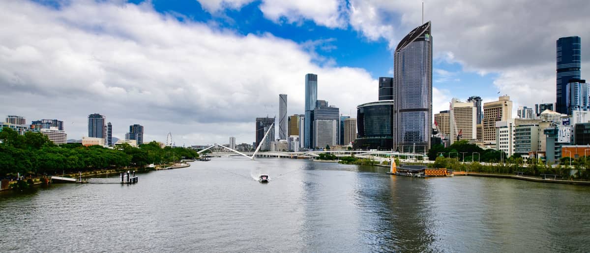 Wide view of Brisbane River with skyline and Victoria Bridge, showing modern city buildings and subtropical greenery under partly cloudy sky.