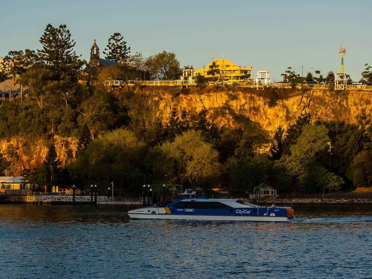 Kangaroo Point cliffs glowing at golden hour above Brisbane River with CityCat ferry passing by, trees and sunlight highlighting the rock face.