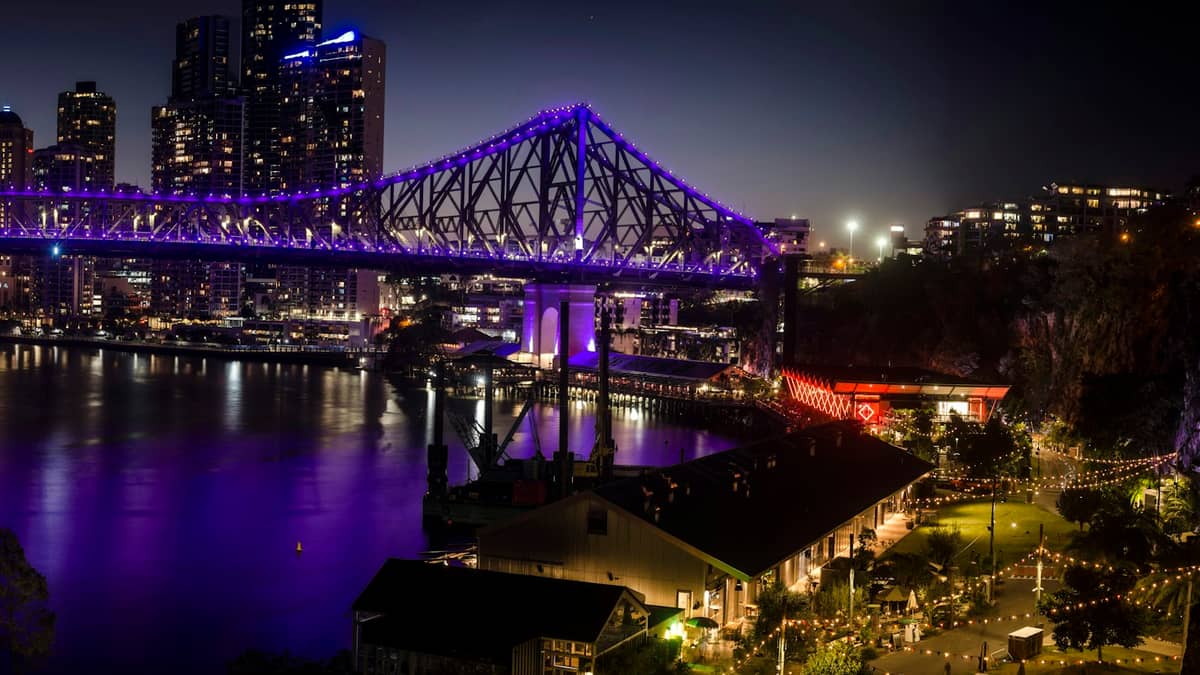 Nighttime view of Brisbane’s Story Bridge illuminated in purple, overlooking the Brisbane River, city lights, and waterfront restaurants with a romantic, vibrant atmosphere.