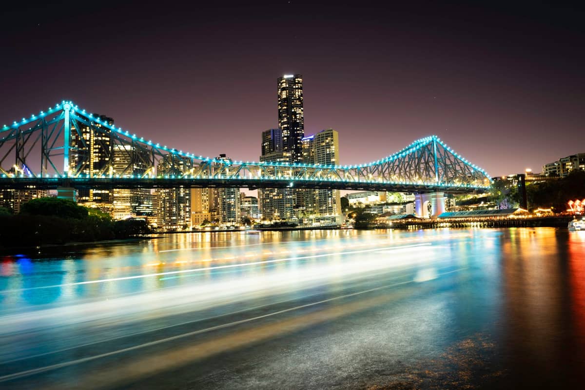 Nachtansicht der Story Bridge in Brisbane, beleuchtet mit blauen Lichtern, mit den Wolkenkratzern der Stadt im Hintergrund und Spiegelungen im Brisbane River.