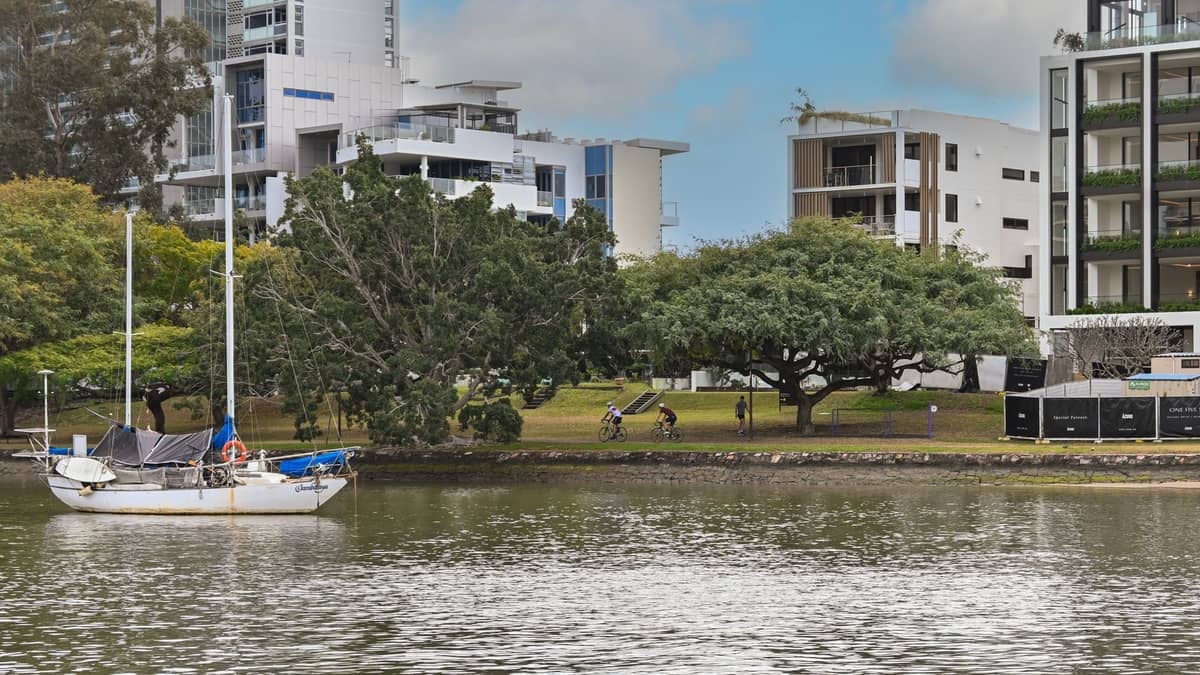 Radfahrer und ein vertäutes Segelboot neben dem New Farm Riverwalk in Brisbane, mit grüner Parkanlage und modernen Apartmentgebäuden im Hintergrund.