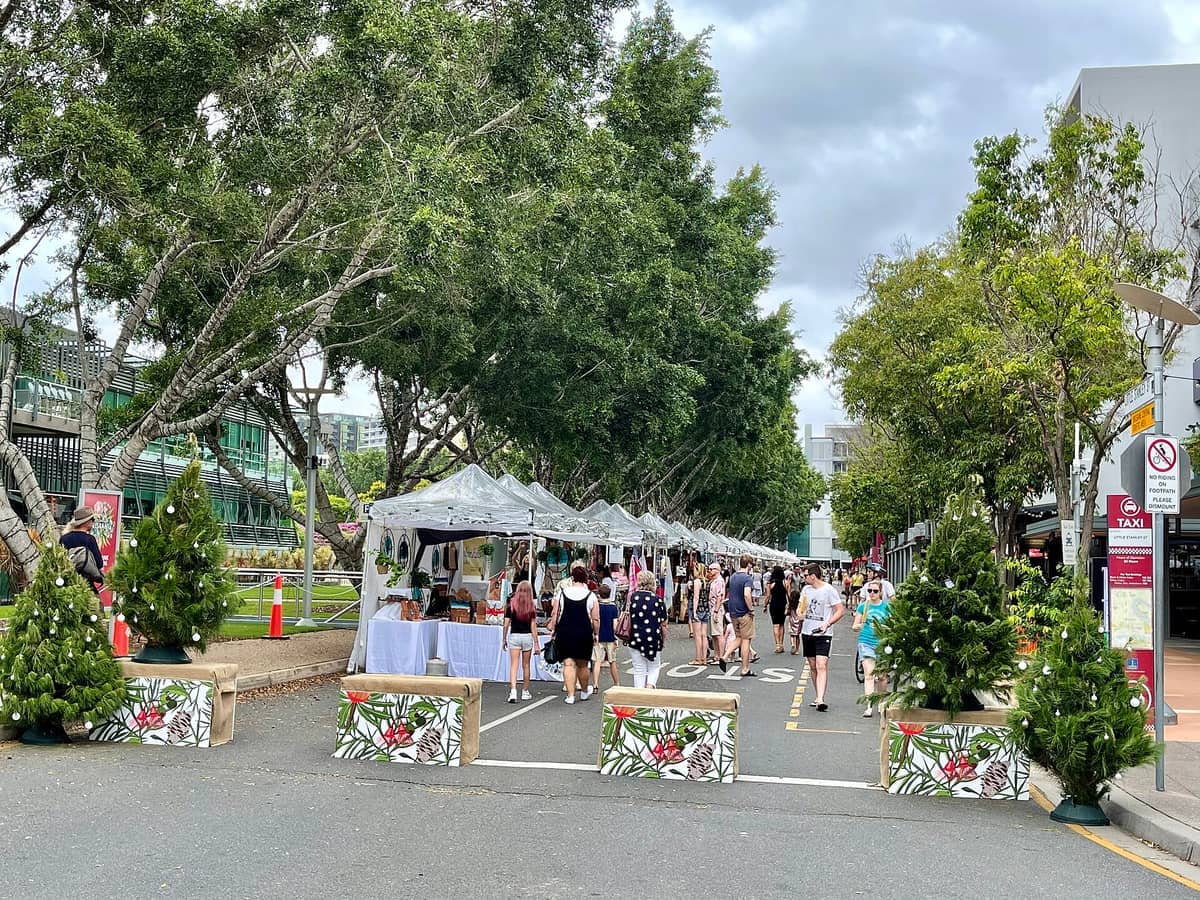 Outdoor-Marktszene in Brisbane mit Reihen weißer Zelte, Einkäufern, die unter großen Bäumen spazieren, und festlichem Grün, das die Straße schmückt.
