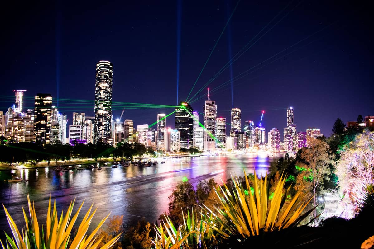Panorama Brisbane nocą z kolorowymi światłami odbijającymi się w rzece i laserami rozświetlającymi niebo — klimat pełen energii i życia nocnego.