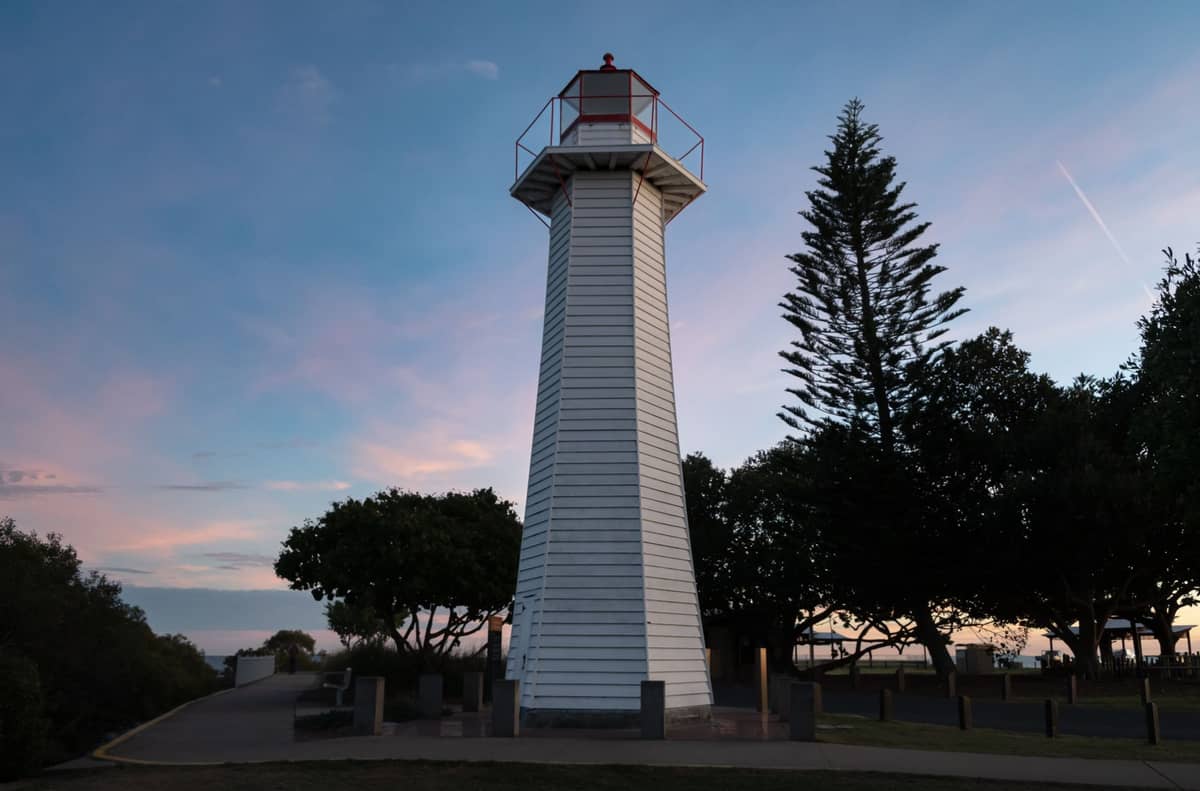 Tall white wooden lighthouse at sunset, surrounded by trees with a path in front, in a coastal park setting.