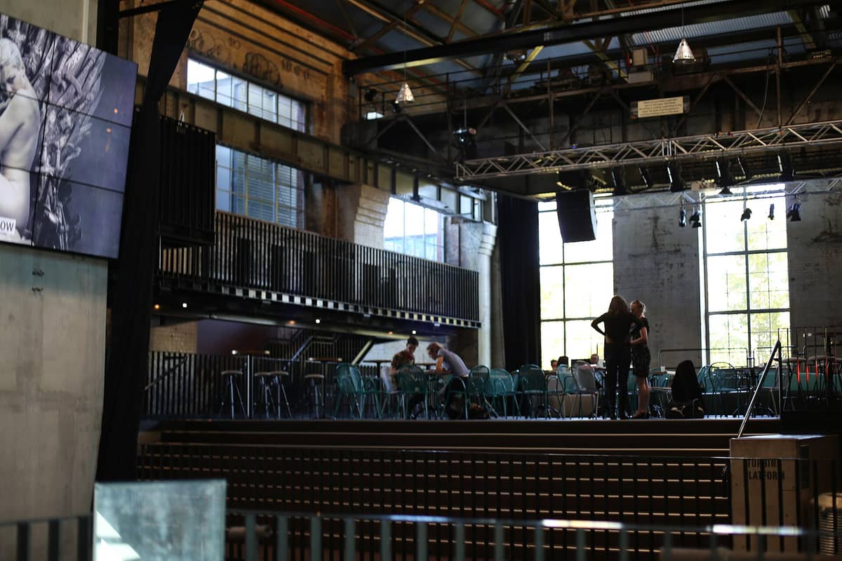 Interior of Brisbane Powerhouse showing exposed brick walls, steel beams, and large industrial windows with people gathered near tables and seating.