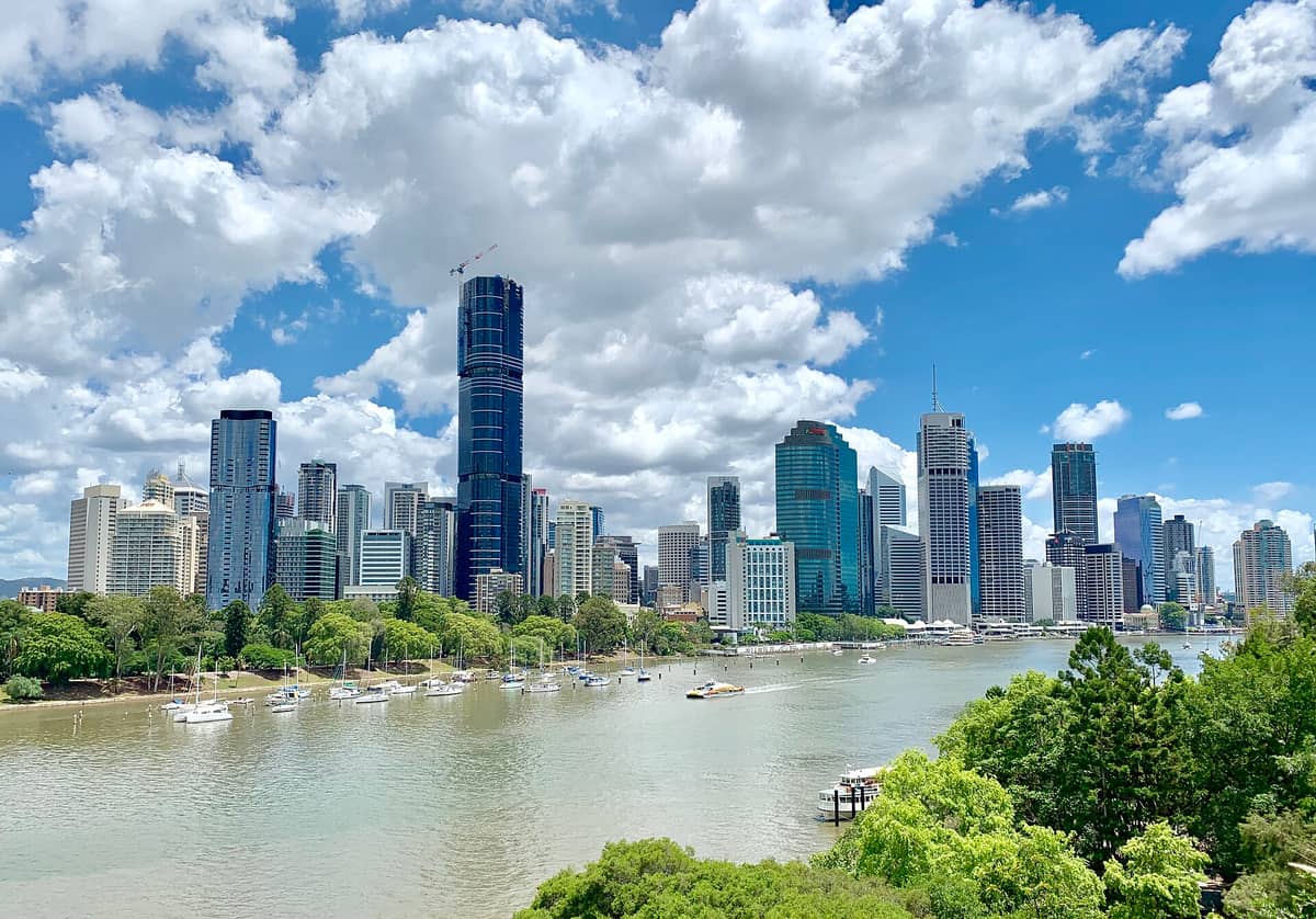 Brisbane River mit festgemachten Segelbooten entlang eines grünen Uferparks, dahinter die Skyline der Stadt und üppiges Grün unter einem sonnigen Himmel