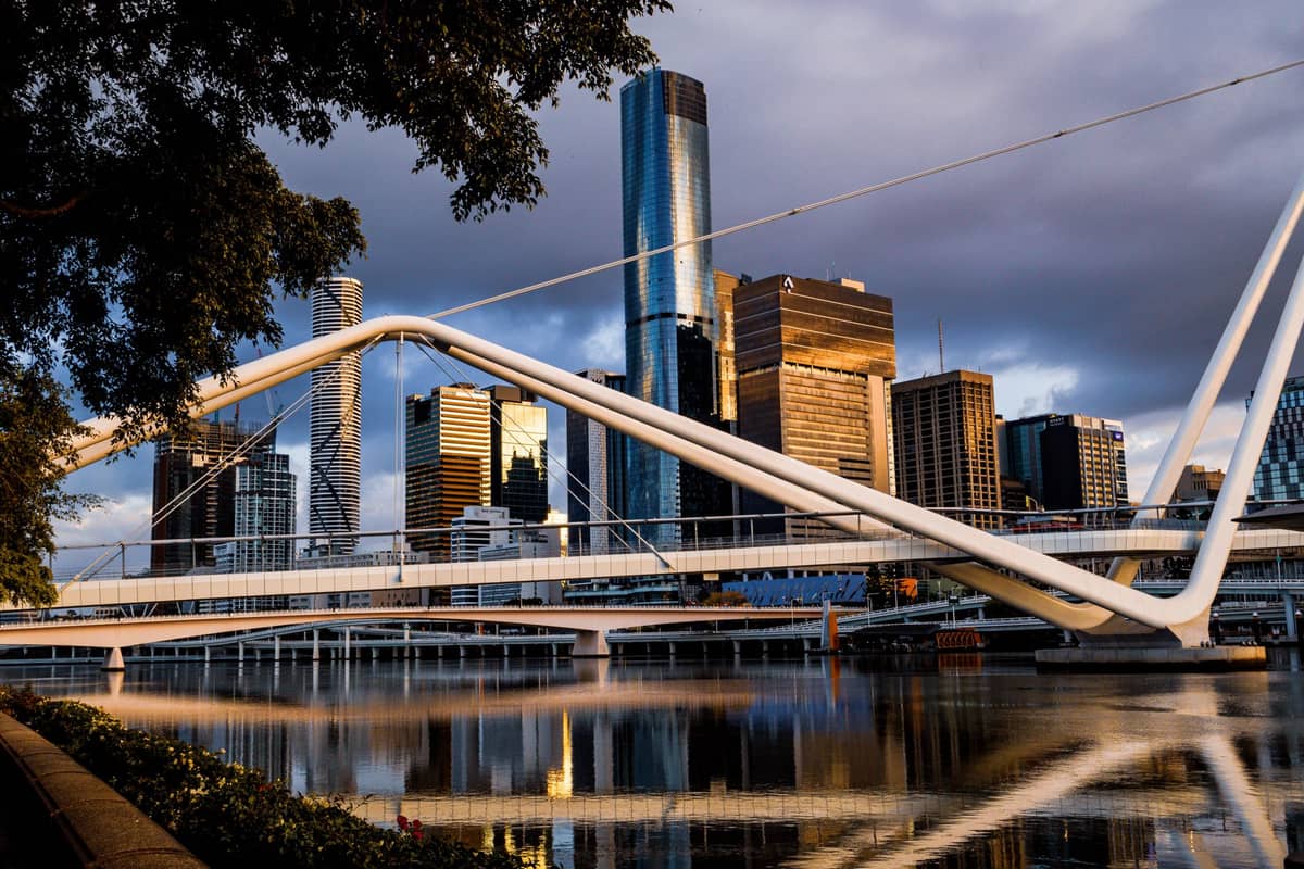 Ein malerischer Uferweg mit der Skyline von Brisbane und einer modernen Fußgängerbrücke, die sich bei Sonnenuntergang im ruhigen Brisbane River spiegeln.