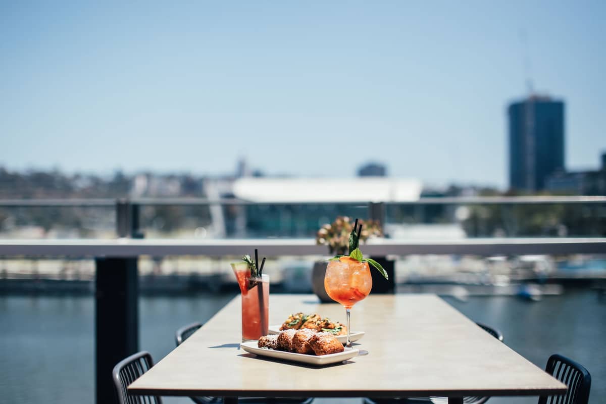 Gedeckter Tisch mit zwei bunten Cocktails und Vorspeisen mit Blick auf den Brisbane River und der verschwommenen Skyline der Stadt im Hintergrund.