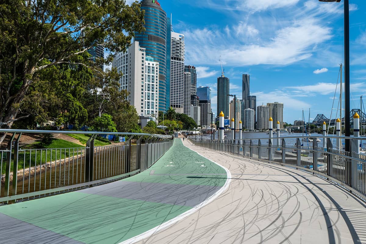 The Brisbane Riverwalk showing dedicated cycling and pedestrian lanes, curving along the river with city skyscrapers and the Story Bridge in the background on a sunny day.