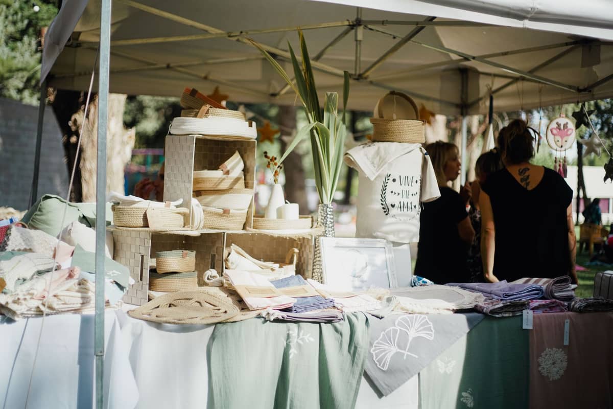 Marktstand im Freien mit verschiedenen lokalen Textilien, handgefertigten Kunsthandwerken und Körben, mit Käufern, die unter einem Sonnendach stöbern.