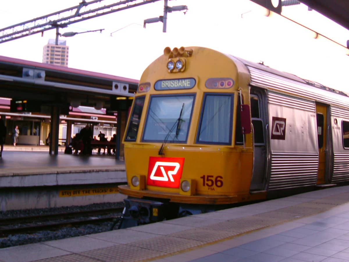 Ein Queensland Rail Zug mit der Aufschrift Brisbane an einem Stadtbahnsteig mit sitzenden und wartenden Fahrgästen in der Nähe.