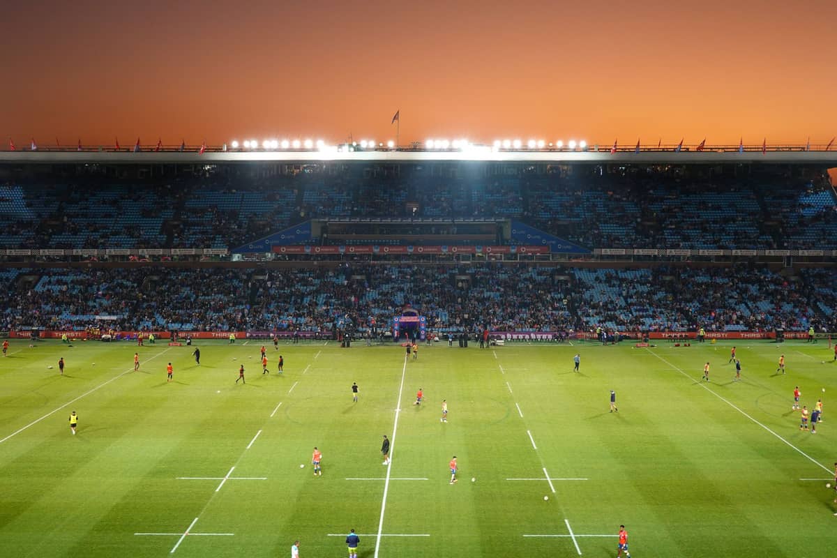 Panoramiczny widok na duży stadion o zachodzie słońca — zawodnicy rozgrzewają się na boisku do rugby, a trybuny wypełniają się kibicami.
