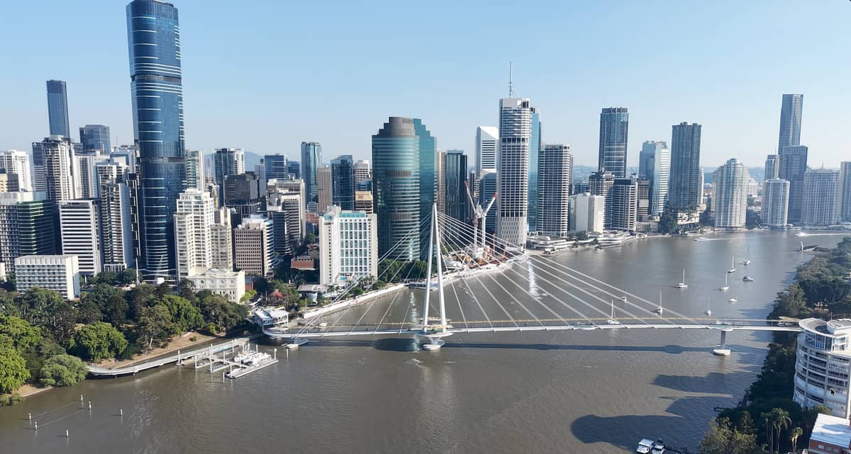 Weitläufiger Blick auf die Skyline von Brisbane mit dem Fluss und der markanten Fußgängerbrücke an einem klaren Tag.