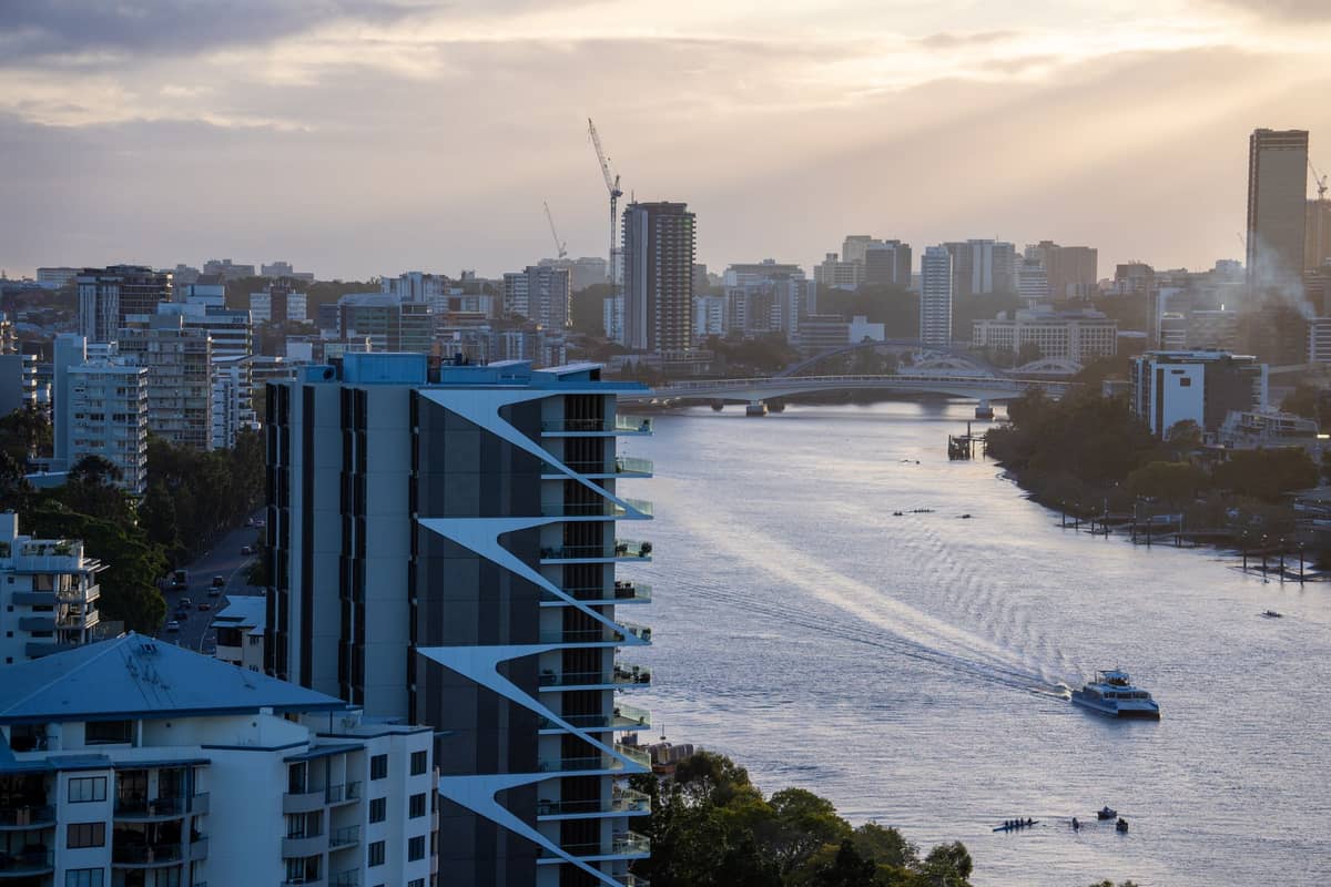 Brisbane River und Stadtpanorama bei Sonnenuntergang mit modernen Gebäuden am Flussufer, fernen Brücken, Booten auf dem Wasser und einem teilweise bewölkten Himmel.