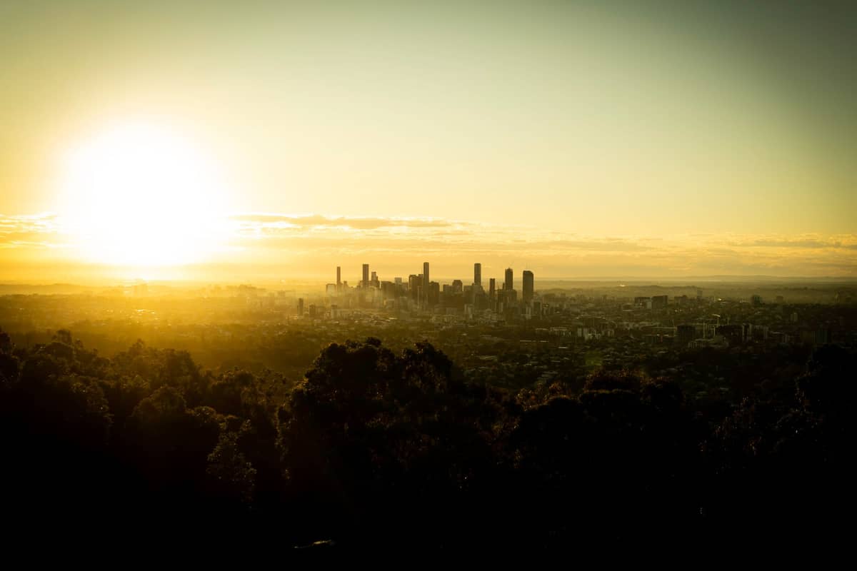 Panorama Brisbane o wschodzie słońca — sylwetki wieżowców na tle nieba, w oddali zarośla Mount Coot-tha i wschodzące słońce nad horyzontem.
