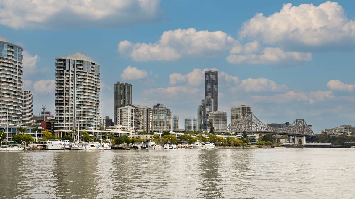 Panorama Brisbane z nowoczesnymi wieżowcami i kultowym mostem Story Bridge w słoneczny dzień, widziana zza rzeki z zacumowanymi łodziami przy nabrzeżu.