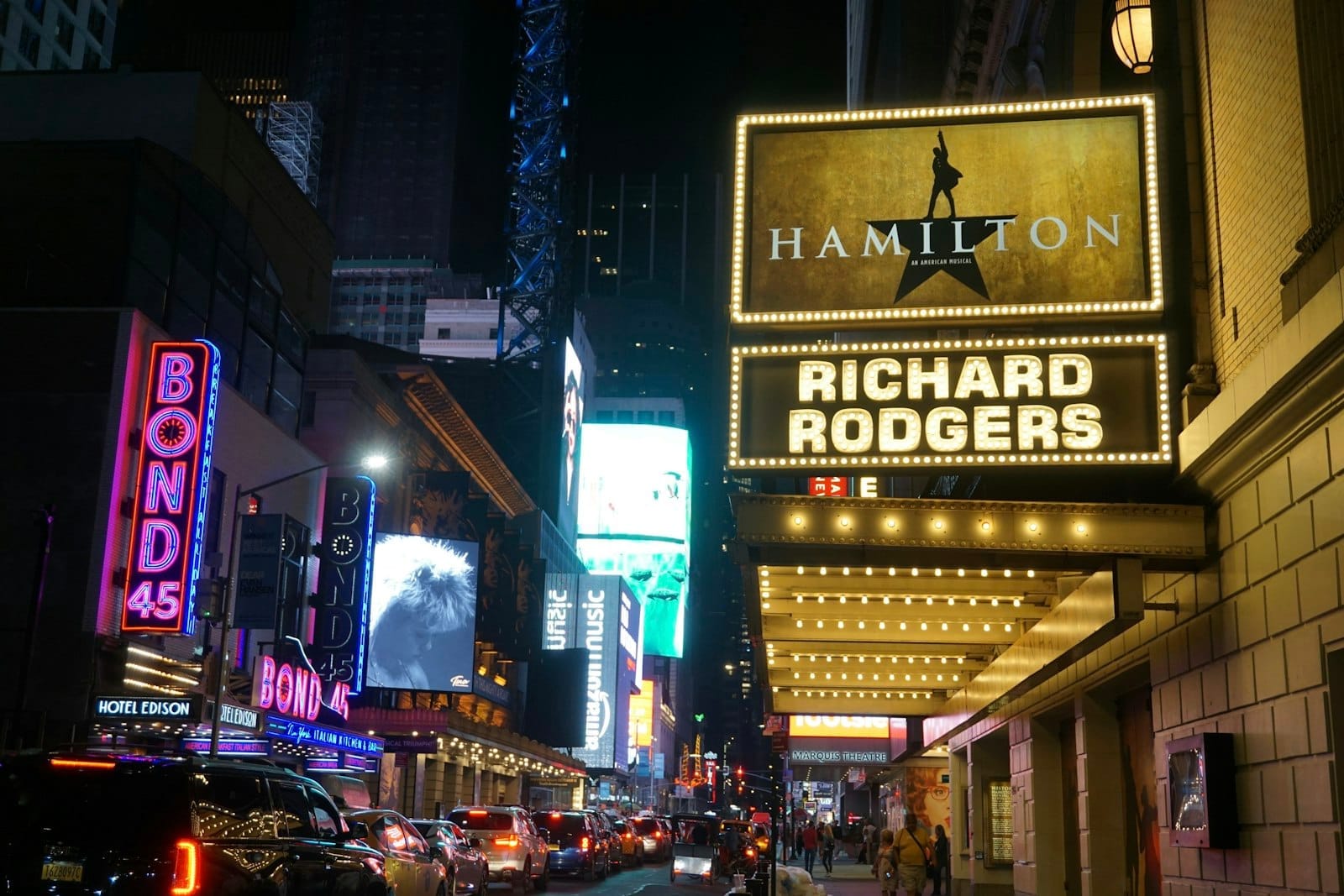 Vista noturna de um teatro da Broadway com o letreiro iluminado de Hamilton no Richard Rodgers Theatre e multidões e carros numa movimentada rua de Nova York.