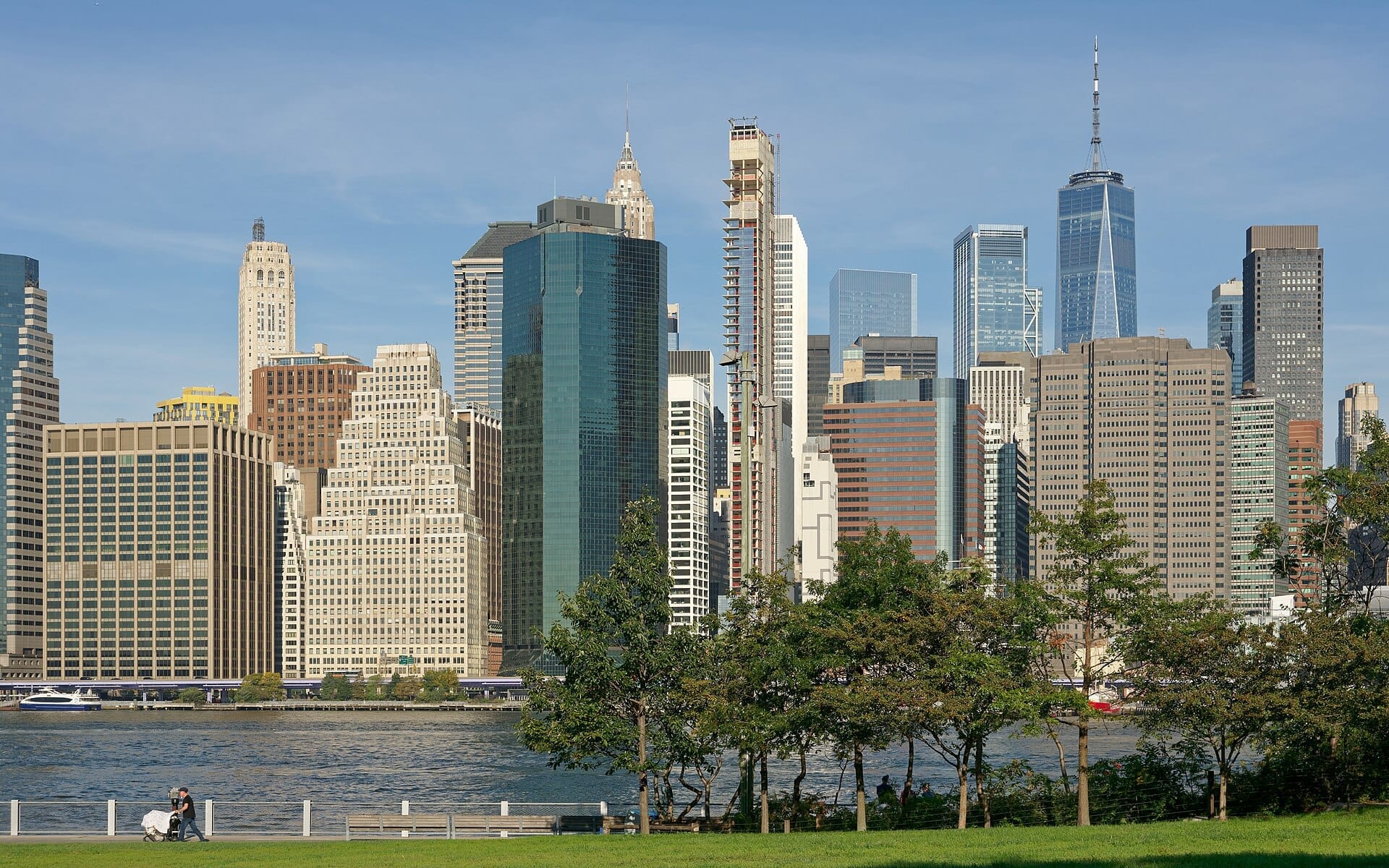 Vista dal Brooklyn Bridge Park con prati verdi, alberi e lo skyline di Manhattan con grattacieli dall'altro lato dell'East River sotto un cielo azzurro.