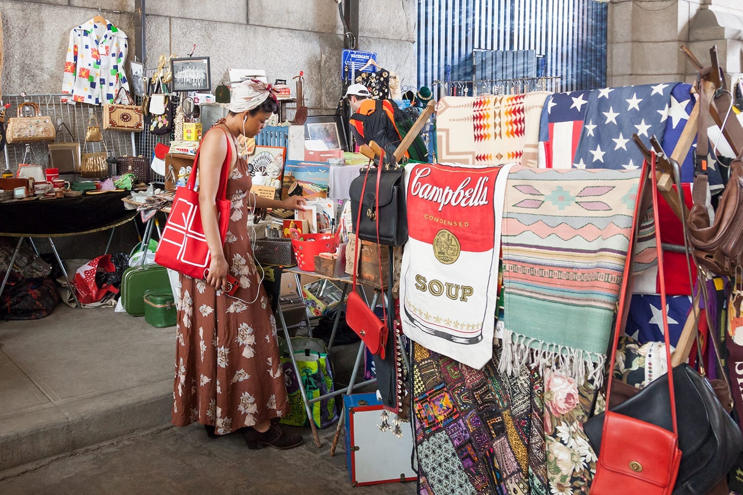 Una donna sfoglia borse vintage e tessuti al Brooklyn Flea DUMBO, con coperte colorate, decorazioni retrò e bancarelle del mercato sotto un arco urbano.