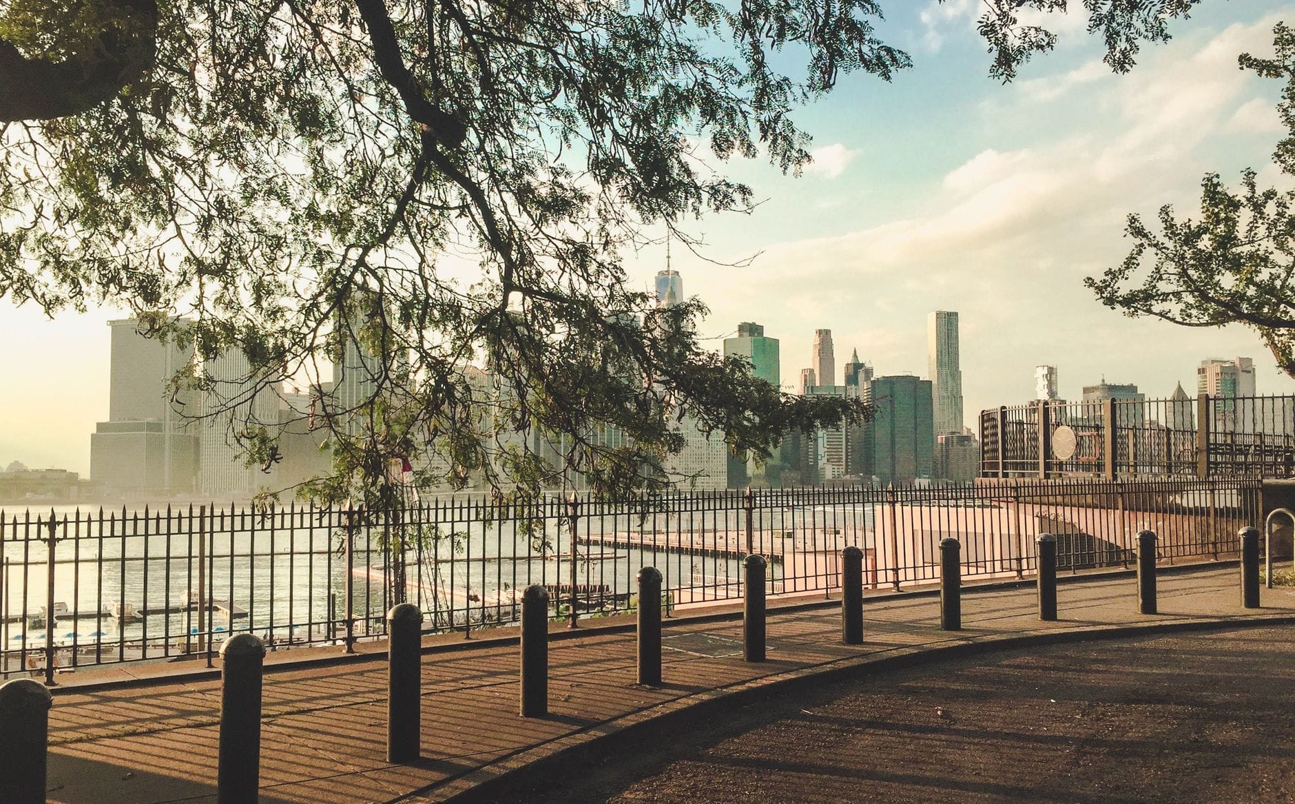 Vista dalla Passeggiata di Brooklyn Heights con lo skyline di Manhattan, il Porto di New York e il Ponte di Brooklyn, incorniciati da alberi illuminati dal sole lungo l'area di osservazione sul lungofiume.