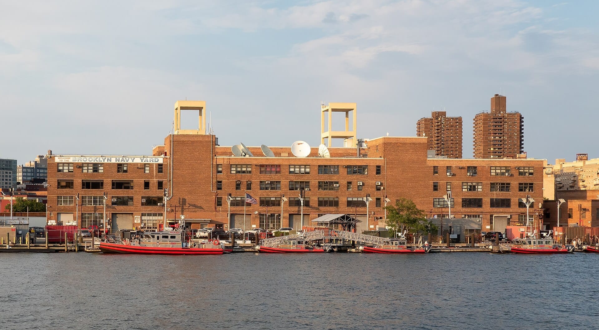Lungomare industriale del Brooklyn Navy Yard con edifici in mattoni, antenne paraboliche, barche rosse ormeggiate e grattacieli della città sullo sfondo in una giornata di sole.