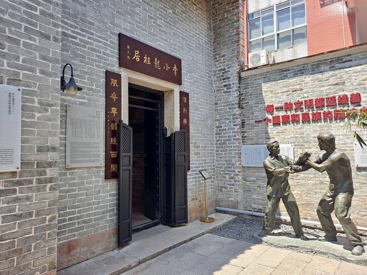 Entrance to Bruce Lee Ancestral Home with Chinese signage, brick walls, and a bronze statue of two martial artists in sunlight.