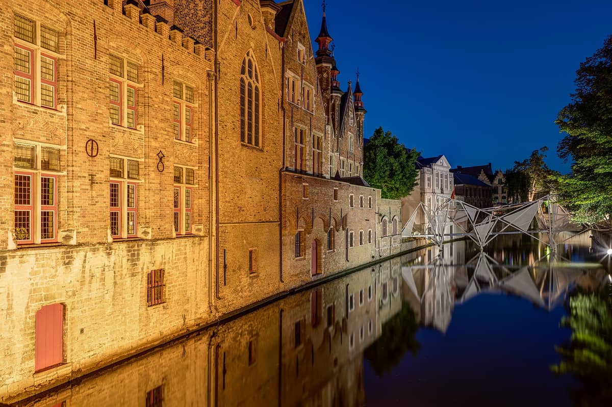 Nighttime view of Bruges canal with illuminated historic brick buildings and modern footbridge reflected in the water, under a deep blue sky.