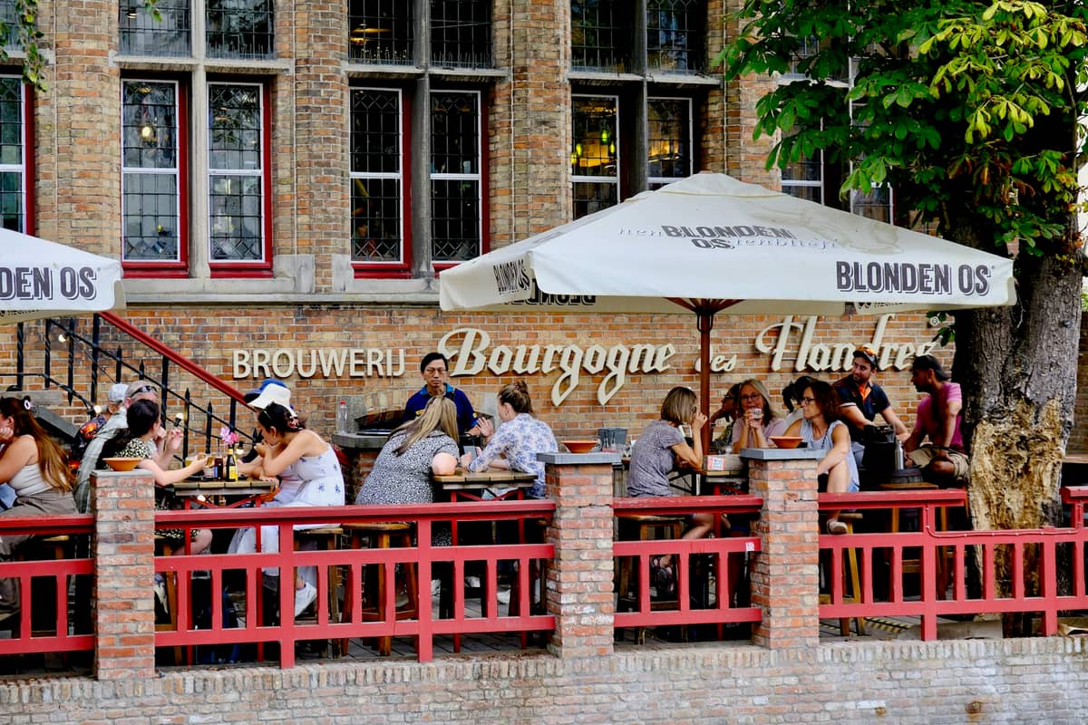 Outdoor terrace at Brouwerij Bourgogne des Flandres in Bruges with people enjoying Belgian beer under branded umbrellas against a historic brick building.