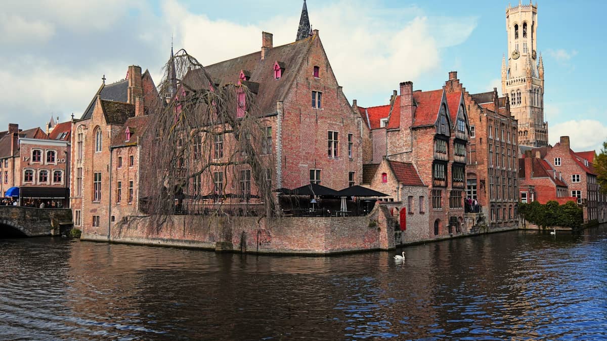 A Bruges canal with an open sightseeing boat cruising past medieval brick buildings and the Belfry tower in the background.