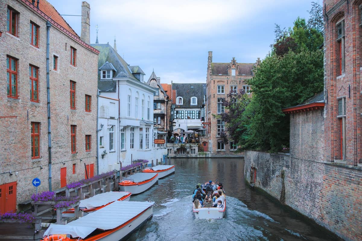 A Bruges canal with a tour boat full of people gliding past charming historic buildings, with more empty tour boats docked along the water's edge.