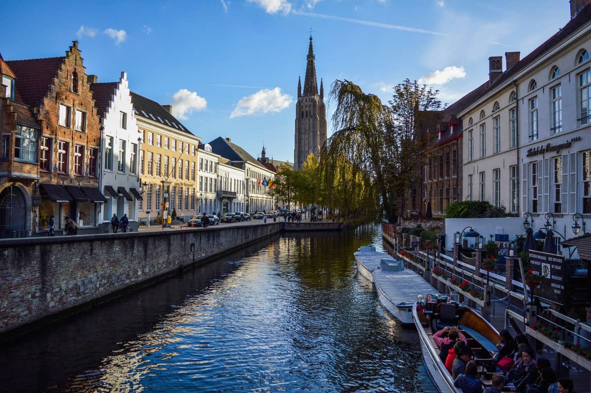 Tour boat with passengers travels along Bruges canal lined by historic buildings, with Church of Our Lady tower visible under blue sky.
