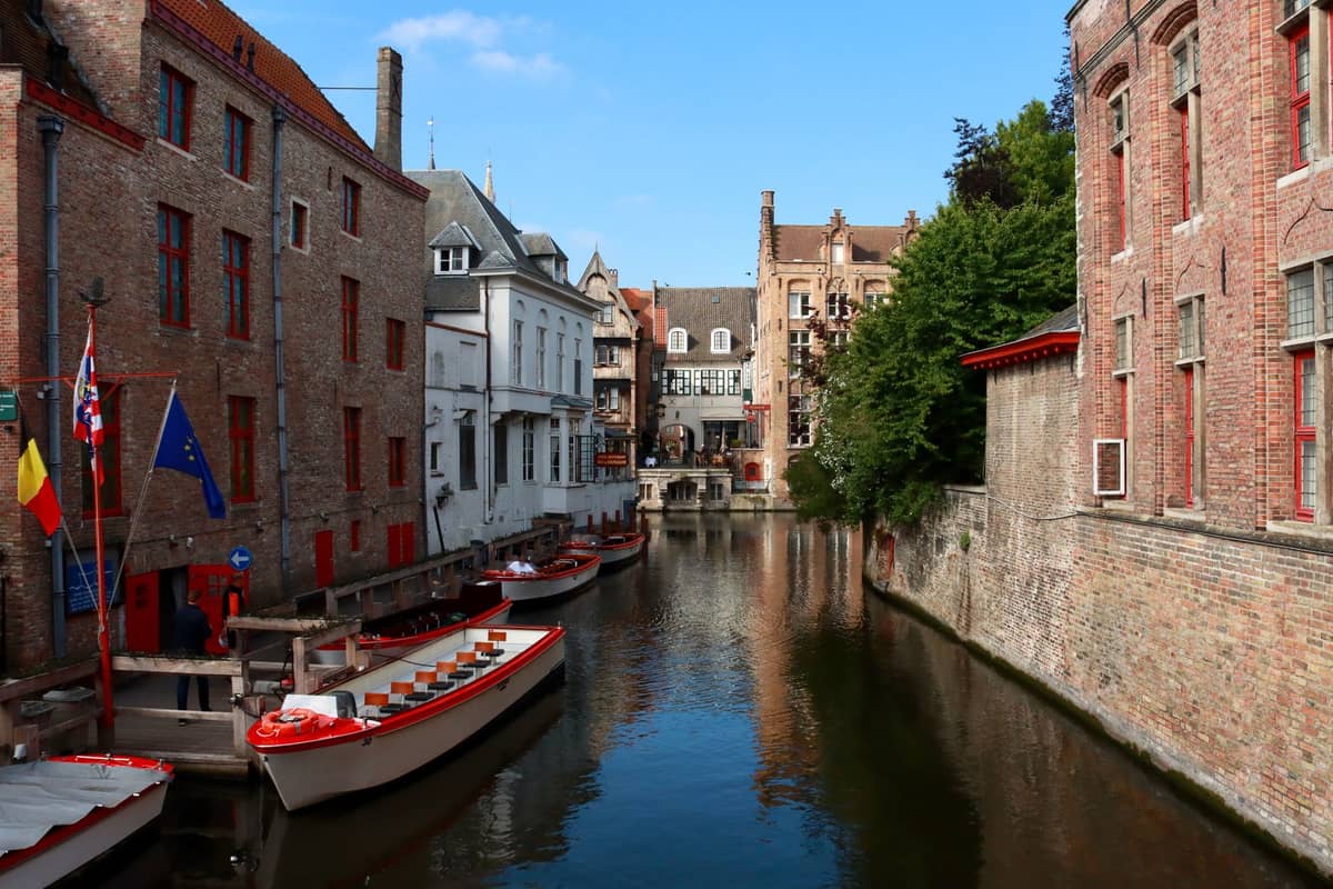 Tour boats docked alongside a canal with historic brick buildings and flags in central Bruges, ready to board visitors.