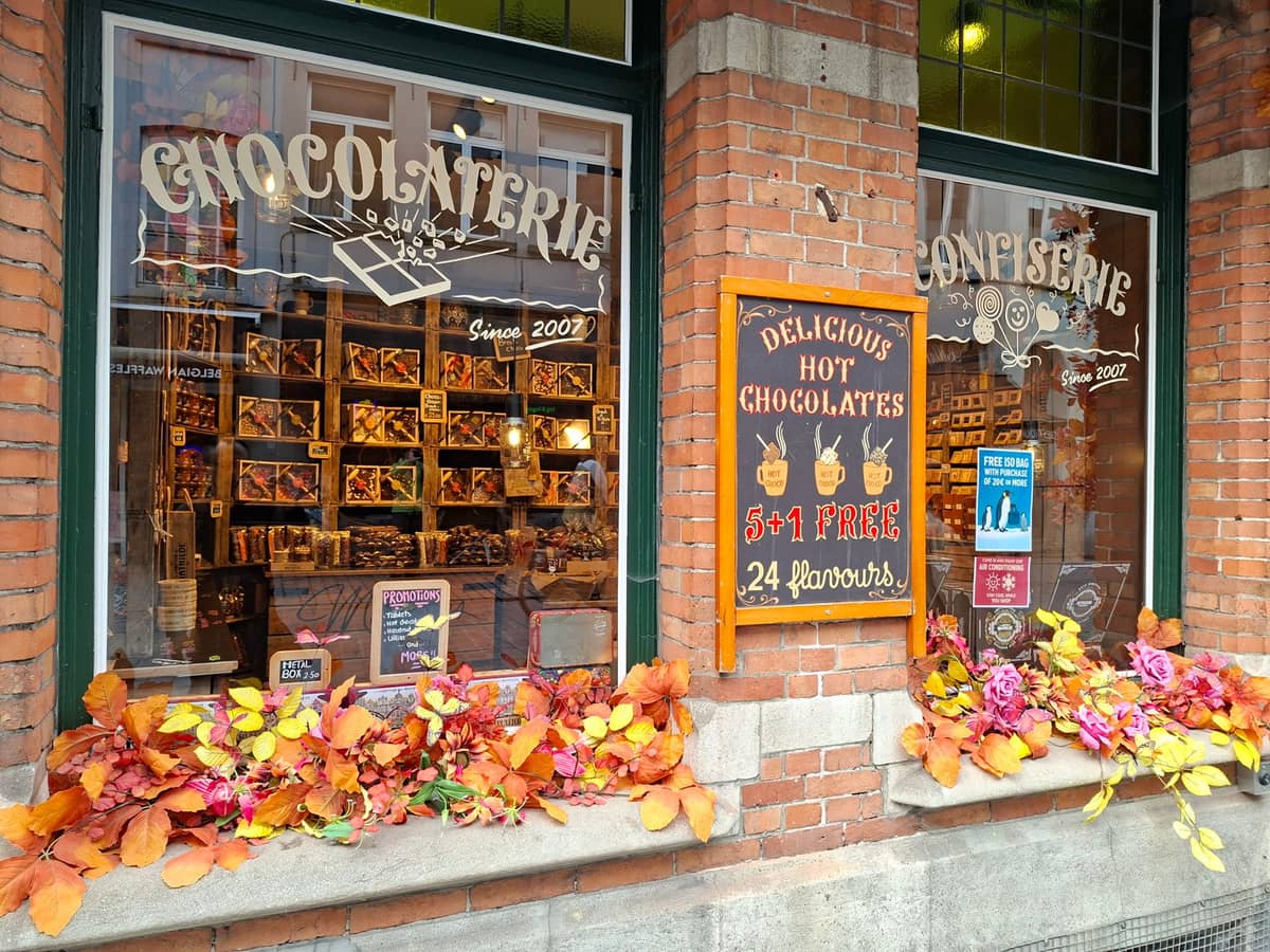 Shopfront of a Bruges chocolaterie with autumn decorations, a window full of artisanal chocolates, and signs advertising hot chocolate and confectionery.