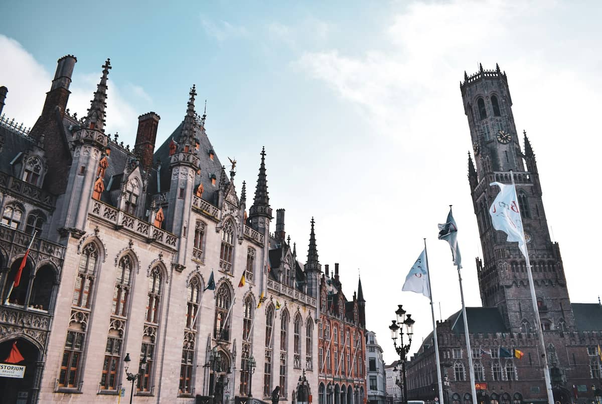 View of Bruges central square Markt with historic guild houses, flagpoles, and the Belfort tower under a bright sky.