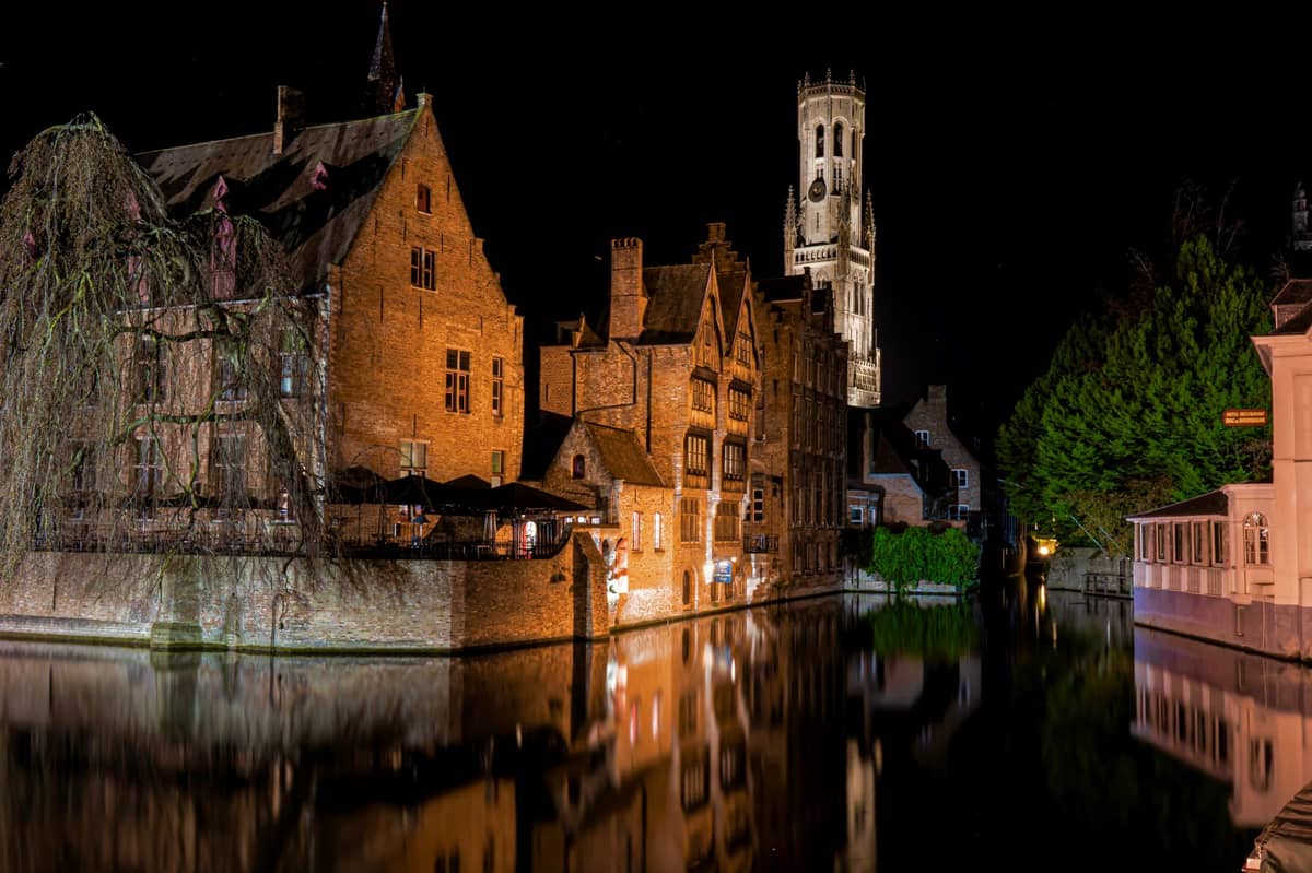 Night view of Bruges canal with medieval buildings illuminated, Belfry tower in the background, and glowing reflections in the water.